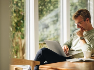  Young man in glasses and a light sweater working happily on a laptop at a wooden table in a bright home office, with large windows...