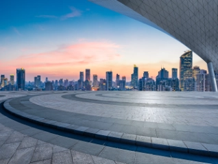 Modern city skyline with high-rises at dusk, viewed from an empty, patterned plaza. A large, curved roof element frames the upper right.