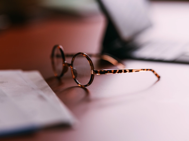 A pair of round, tortoise-shell eyeglasses rests on a desk next to a sheet of paper, with a soft, warm light casting a shallow depth of field.