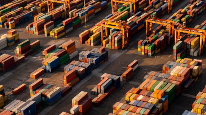 Aerial view: Hundreds of brightly colored shipping containers are stacked high at a busy port/terminal, with large yellow gantry cranes overhead.
