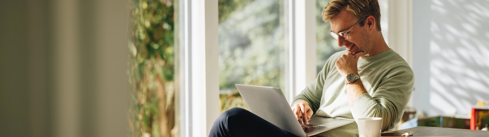  Young man in glasses and a light sweater working happily on a laptop at a wooden table in a bright home office, with large windows overlooking greenery.