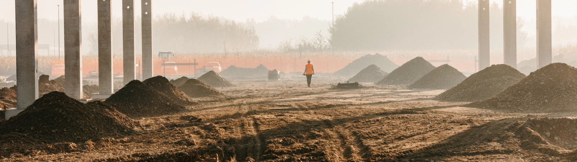 Worker in orange vest walks across muddy construction site with dirt mounds, framed by concrete pillars & misty sun.