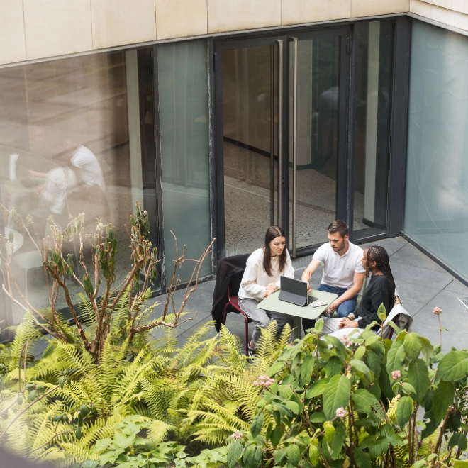 Two women, one man sit at a small outdoor table, looking at a laptop. Green foliage is in the foreground, large glass doors behind.